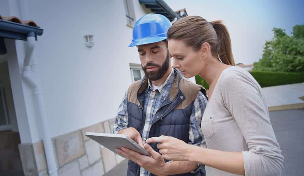Male service technician wearing a hard hat seen from the waist up, showing a female customer information on a tablet or digital device