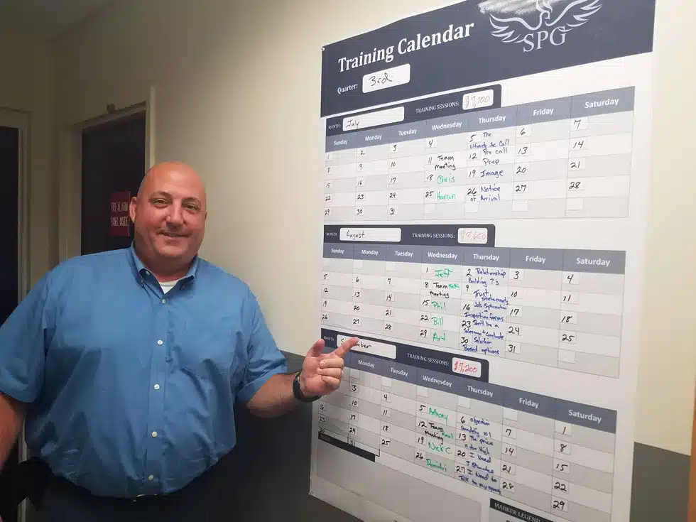 Male business owner wearing a blue dress shirt, smiling and standing in front of and pointing at a Service Profit Group Training calendar wall display