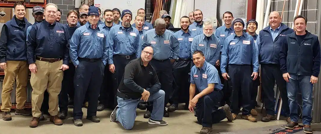 Group photo of technicians, owners, and Joe O'Grady kneeling in front of group, most wearing uniforms in warehouse